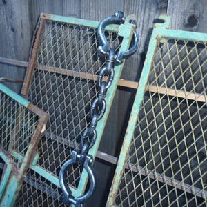 Op de afbeelding: Een zwarte metalen ketting met twee handboeien aan de uiteinden. De ketting hangt aan een groen metalen hek met een ruitvormig patroon.