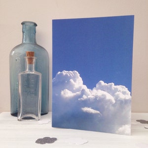 Może przedstawiać: Kartka z pozdrowieniami z motywem błękitnego nieba i chmur stoi obok dwóch szklanych butelek. Jedna butelka jest przezroczysta z korkiem, a druga to duża, niebieska, antyczna butelka. Konfetti w kształcie chmur jest rozrzucone.