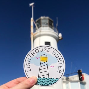 May include: A white sticker with a blue, yellow, and white lighthouse illustration and the text "LIGHTHOUSE HUNTER" in a circle. The sticker is being held in front of a white lighthouse.