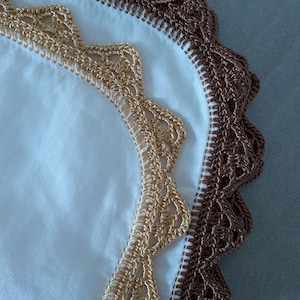May include: A white fabric textile featuring two distinct layers of decorative crochet edging. The inner border is crafted from metallic gold thread in a scalloped pattern, while the outer border consists of dark brown thread in a similar design.