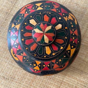 May include: A round wooden box with a lid featuring a black base and intricate geometric patterns in red, yellow, and green. The design consists of a central star-shaped motif surrounded by concentric circles of repeating triangular and petal shapes.