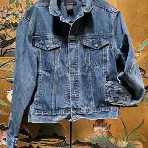 May include: A medium-wash blue denim jacket featuring a classic collar, long sleeves with buttoned cuffs, and two chest pockets with button-down flaps. The jacket includes a front button closure and a Calvin Klein brand label at the inner neckline.