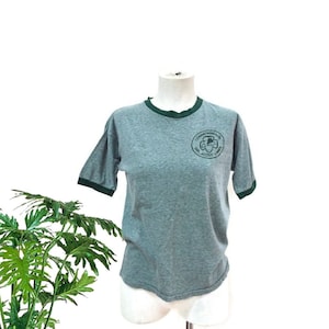 May include: Grey ringer t-shirt with dark green trim on the collar and sleeves. The shirt has a circular emblem on the chest that reads "Commonwealth Girl Scout Camp" in green lettering. The shirt is displayed on a mannequin.