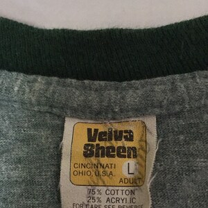 May include: Close-up of a grey t-shirt collar with a dark green trim. A yellowed tag reads "Velva Sheen" and "Cincinnati, Ohio, U.S.A." with size "L" and fabric content: 75% cotton, 25% acrylic.