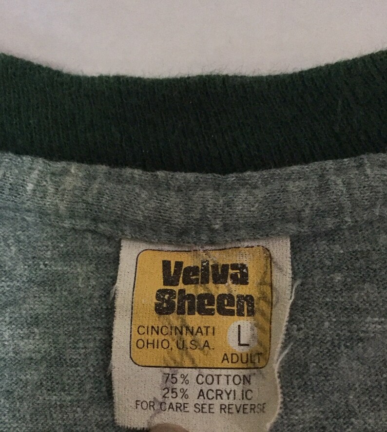 May include: Close-up of a grey t-shirt collar with a dark green trim. A yellowed tag reads "Velva Sheen" and "Cincinnati, Ohio, U.S.A." with size "L" and fabric content: 75% cotton, 25% acrylic.