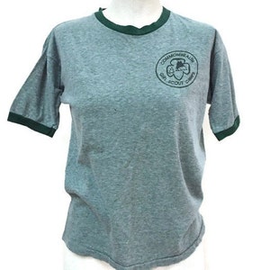 May include: Grey t-shirt with green trim on the collar and sleeves. The shirt features a circular emblem with the text "Commonwealth Girl Scout Camps". The t-shirt is displayed on a mannequin.