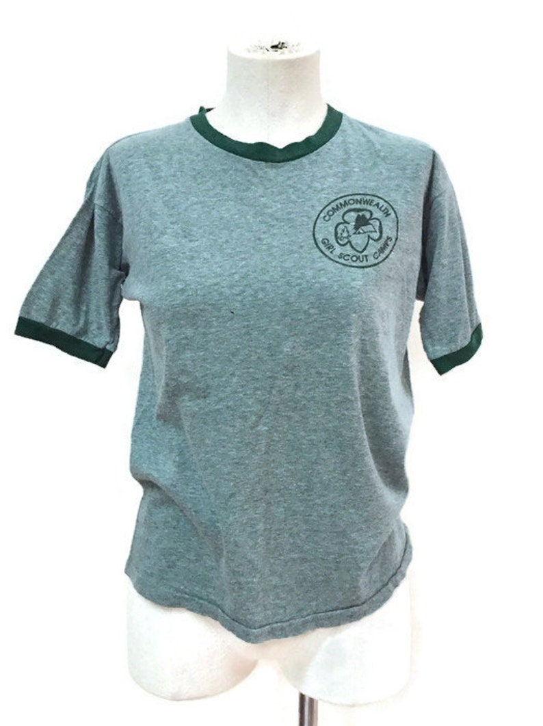 May include: Grey t-shirt with green trim on the collar and sleeves. The shirt features a circular emblem with the text "Commonwealth Girl Scout Camps". The t-shirt is displayed on a mannequin.