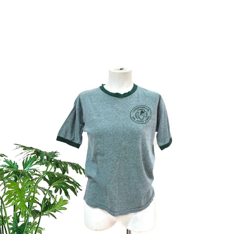 May include: Grey ringer t-shirt with dark green trim on the collar and sleeves. The shirt has a circular emblem on the chest that reads "Commonwealth Girl Scout Camp" in green lettering. The shirt is displayed on a mannequin.