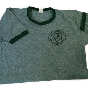 May include: A grey t-shirt with dark green trim around the collar and sleeves. The shirt has a circular emblem with the text "Commonwealth Girl Scout Camps". The t-shirt is made of a soft material, possibly cotton.