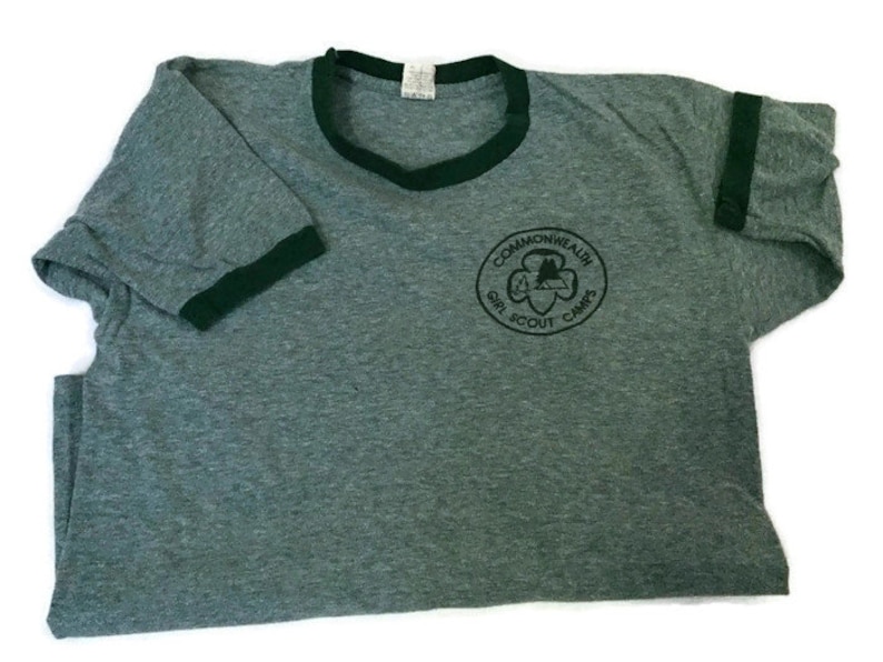 May include: A grey t-shirt with dark green trim around the collar and sleeves. The shirt has a circular emblem with the text "Commonwealth Girl Scout Camps". The t-shirt is made of a soft material, possibly cotton.