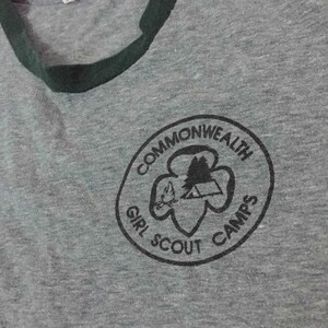 May include: Grey t-shirt with a dark green collar and trim. The shirt has a circular graphic with the words "Commonwealth Girl Scout Camps" around a design of a tent, trees, and a flame.