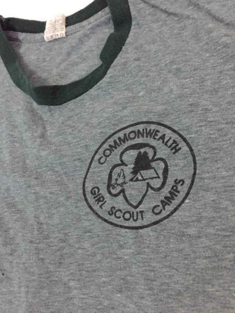 May include: Grey t-shirt with a dark green collar and trim. The shirt has a circular graphic with the words "Commonwealth Girl Scout Camps" around a design of a tent, trees, and a flame.