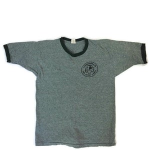 May include: Grey ringer t-shirt with a black collar and sleeve cuffs. The shirt has a circular graphic on the front with the text "Commonwealth Girl Scout Camp". The t-shirt is made of a soft, worn material.