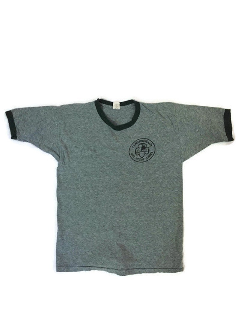 May include: Grey ringer t-shirt with a black collar and sleeve cuffs. The shirt has a circular graphic on the front with the text "Commonwealth Girl Scout Camp". The t-shirt is made of a soft, worn material.