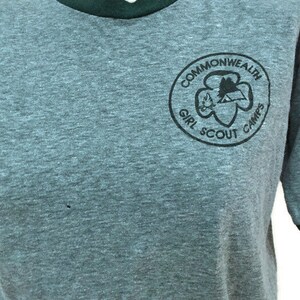 May include: A grey t-shirt with a dark green collar and sleeve trim. The shirt displays a circular emblem with the words "Commonwealth Girl Scout Camps" in black. The emblem includes a trefoil and a mountain scene.