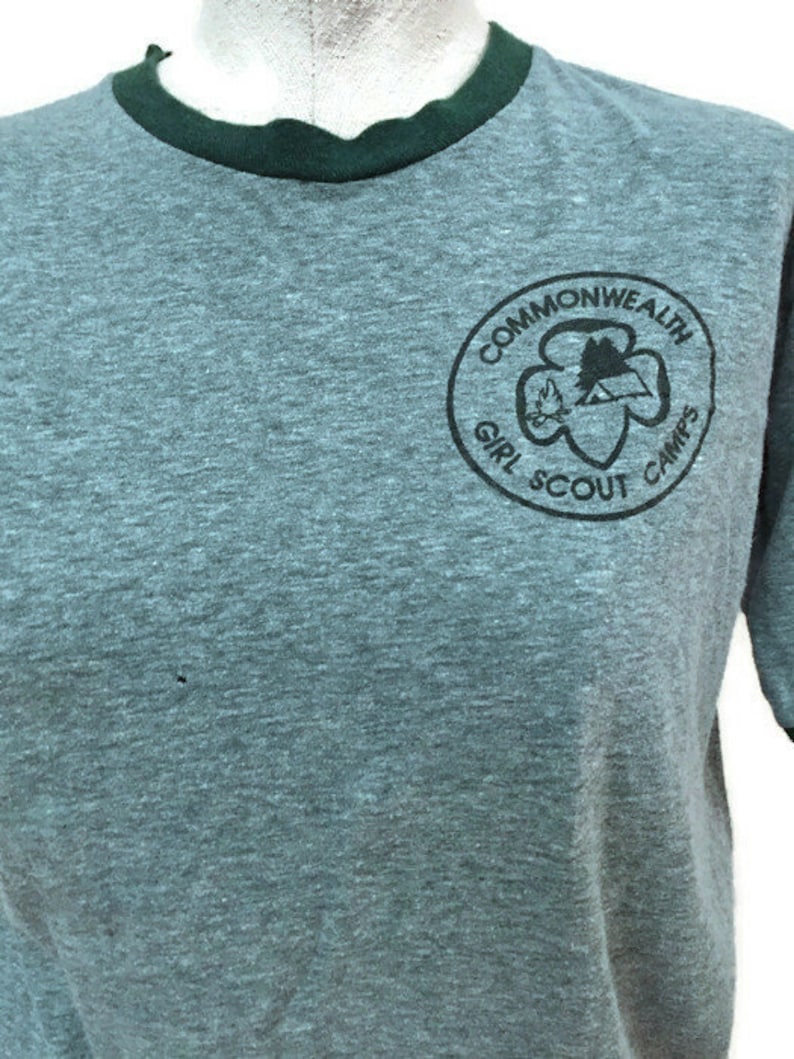 May include: A grey t-shirt with a dark green collar and sleeve trim. The shirt displays a circular emblem with the words "Commonwealth Girl Scout Camps" in black. The emblem includes a trefoil and a mountain scene.