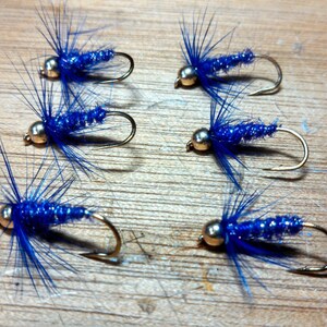 May include: Six blue fishing flies with gold-colored hooks and heads are arranged on a wooden surface. The flies have a vibrant blue body and feathery blue extensions, suggesting they are designed to mimic insects.