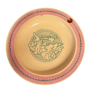 Può includere: Un piatto per bambini vintage con interno giallo chiaro e bordo rosa. Il piatto presenta un'illustrazione verde di una cicogna che trasporta un bambino. C'è un piccolo foro sul bordo. Il testo "©1983 OAA, INC." è stampato sul piatto.