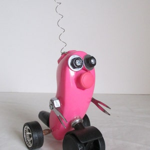 May include: A pink robot toy with a spring antenna and a pink pom-pom on top. The robot has black wheels and is made from recycled materials.