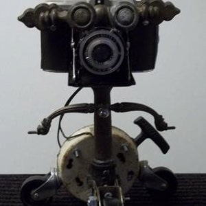 May include: A steampunk-style camera on wheels with a black and silver finish. The camera has a large lens and a tripod-like base with three wheels.
