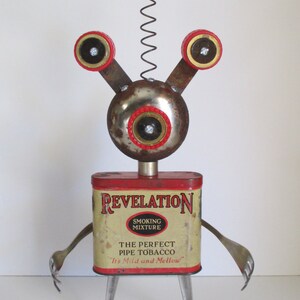 May include: A whimsical robot made from recycled materials, featuring a red and white Spiderman head, red and gold eyes, and a rusty metal body. The robot is standing on two silver spoons and holding a vintage tin can labeled "Revelation Smoking Mixture The Perfect Pipe Tobacco "It's Mild and Mellow."