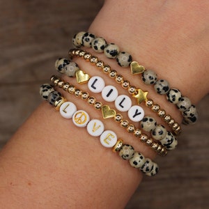 May include: A set of five bracelets with gold beads and dalmatian jasper stones. The bracelets feature gold charms, including hearts, stars, and a peace sign. The bracelets also have white beads with the letters "LOVE" and "LILY" spelled out.