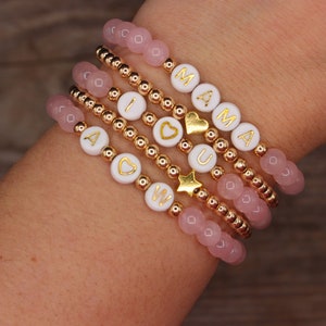 May include: A set of six bracelets with pink and gold beads. The bracelets spell out "I LOVE YOU MOM" with gold letters on white beads. The bracelets are stacked on a person's wrist.