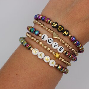 May include: Five bracelets stacked on a wrist. The bracelets are made of small gold and iridescent beads. The bracelets spell out "MAMA", "LOVE", and "MOM" with gold letters on white and black beads.