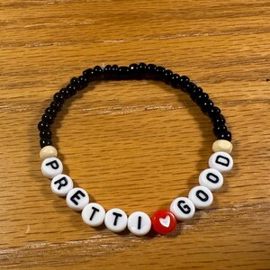May include: A beaded bracelet with black, white, and red beads. The bracelet spells out "PRETTY GOOD" with white letter beads. A red heart bead is between the words. The bracelet is on a wooden surface.