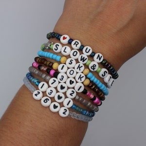 May include: A stack of colorful beaded bracelets with letter and number charms. The bracelets spell out "RUN & SLOW IS OK" and include the numbers 1, 2, 3, 6, and 7.  The bracelets are on a person's wrist.