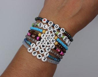 May include: A stack of colorful beaded bracelets with letter and number charms. The bracelets spell out "RUN & SLOW IS OK" and include the numbers 1, 2, 3, 6, and 7.  The bracelets are on a person's wrist.