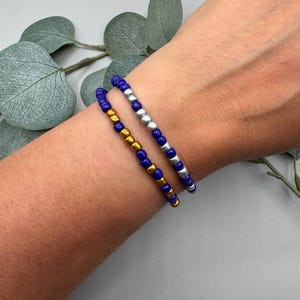 May include: Two beaded bracelets are displayed on a wrist. One bracelet features small, dark blue beads, and the other has silver beads. A second bracelet has gold beads. The bracelets are stacked together. Green leaves are in the background.