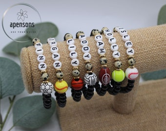 May include: Seven personalized bracelets with white beads and black letters spelling out the names Finn,  Organ, Lily, Addy, Cam, Holly, and  Esley. Each bracelet has a black elastic band and a sports ball charm: soccer, softball, basketball, football, tennis ball, and baseball.