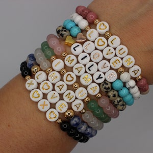 May include: A stack of bracelets with white beads and gold lettering spelling out words like "love", "luna", and "mama". The bracelets are made with different colored stones, including turquoise, pink, and green.