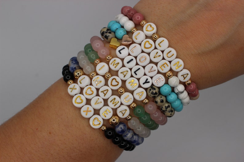 May include: A stack of bracelets with white beads and gold lettering spelling out words like "love", "luna", and "mama". The bracelets are made with different colored stones, including turquoise, pink, and green.