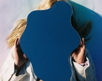 May include: A person with blonde hair is holding a blue, irregularly shaped mirror in front of their face. They are wearing a white blazer and pink pants.
