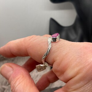 May include: A silver ring with a pink gemstone. The ring features a twisted band design and a rectangular pink stone. The ring is on a finger.