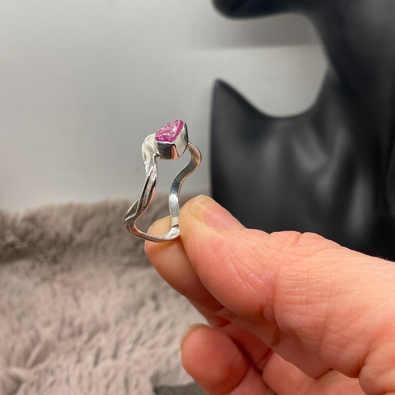 May include: A silver ring featuring a pink, heart-shaped gemstone. The ring has a distinctive, flowing design with a twisted band element. The gemstone is bezel-set. The ring is held in a person's hand.
