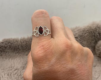 May include: A silver ring featuring a central marquise-cut deep red gemstone. The band is designed with openwork metal detailing that incorporates heart shapes on both sides of the stone, creating a symmetrical and intricate decorative pattern.