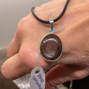 Pode incluir: Pingente oval com uma pedra preciosa de corindo estrela marrom emoldurada em uma armação de metal prateado, suspenso por um cordão preto. O pingente é segurado por uma mão, com uma etiqueta "STAR corundum".