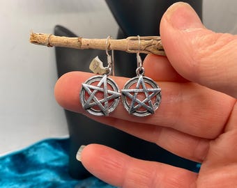 May include: A pair of silver-colored earrings featuring a pentagram design. The earrings are round with a star shape inside a circle. The earrings are held up by a small wooden stick. The background is a dark color.