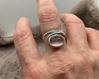 Puede incluir: Un anillo de plata tipo bypass con una piedra cabujón ovalada de color rosa pálido y superficie lisa. La banda está hecha de metal plateado pulido que se envuelve alrededor del dedo, con la piedra engastada en un bisel plateado sencillo.