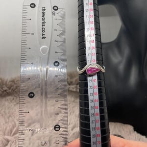 May include: A silver ring with a pink gemstone, shown on a ring sizer. The ring features a wavy design and a triangular pink gemstone. A ruler with cm measurements is visible.