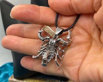 May include: A silver-colored scorpion pendant on a black cord necklace. The scorpion is highly detailed, with visible claws, legs, and body segments. The pendant is held in a hand, with the word "Scorpio" visible on a small tag.