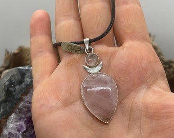 May include: A silver pendant necklace with a pink rose quartz teardrop-shaped stone and a silver crescent moon charm. The necklace is on a brown cord.