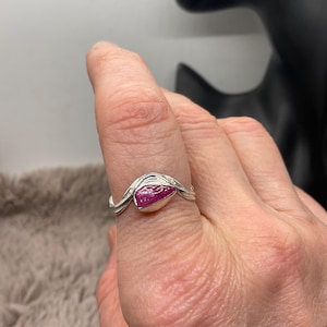 Peut inclure: Une bague en argent ornée d'une pierre précieuse rouge rubis de forme ovale. L'anneau présente un motif tourbillonnant délicat avec de petites pierres d'accent claires. La pierre précieuse est au centre, sertie dans un sertissage en argent poli.