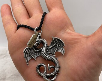 Può includere: Un ciondolo a forma di drago argentato con dettagli intricati, tra cui ali e squame, è appeso a un cordino nero. Una piccola etichetta è attaccata al cordino. Il ciondolo è tenuto in una mano aperta.