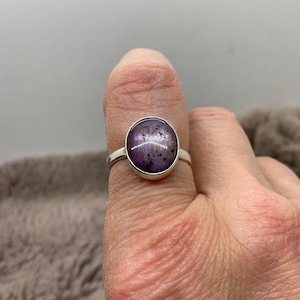 以下が含まれることがあります： 大きな丸い紫色の宝石と暗いインクルージョンが付いたシルバーリング。リングは、シンプルで磨かれたシルバーバンドにセットされています。宝石は、磨かれた滑らかな表面を持っています。