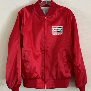 May include: A red satin bomber jacket with a zipper closure and ribbed cuffs. The jacket has a Champion Truck Plug logo on the left chest.