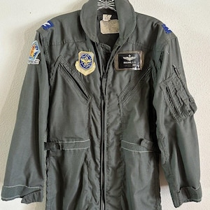 May include: A dark green flight suit with multiple patches, including a US Air Force patch, a name tag that reads "BULLARD, J.S.", and a patch with a globe and wings. The suit has a zipper closure and multiple pockets.